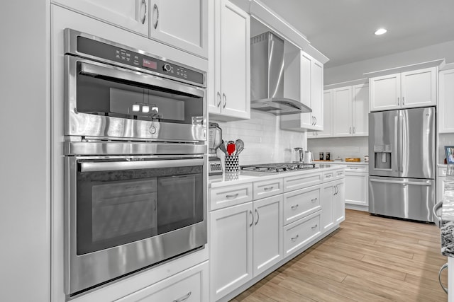 kitchen with white cabinets, wall chimney range hood, stainless steel appliances, tasteful backsplash, and light wood-type flooring