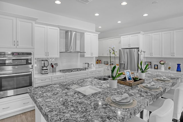 kitchen featuring wall chimney exhaust hood, white cabinetry, stainless steel appliances, an island with sink, and a breakfast bar area