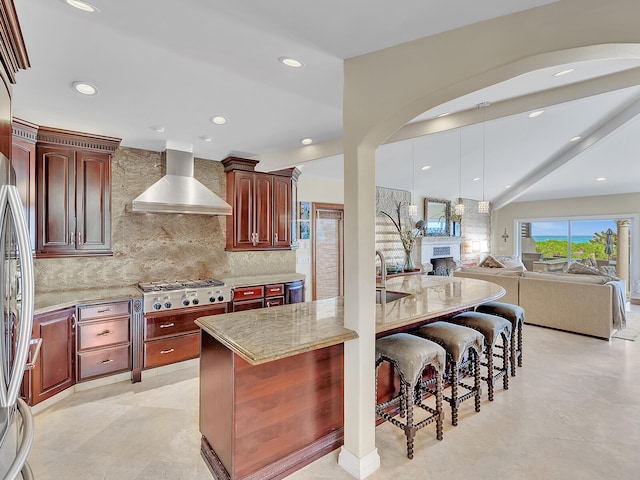 kitchen featuring light stone countertops, wall chimney range hood, a breakfast bar area, stainless steel gas cooktop, and a center island with sink