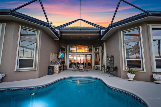 pool at dusk with glass enclosure, ceiling fan, and a patio area