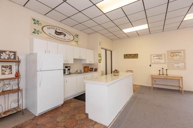 kitchen with a drop ceiling, freestanding refrigerator, light countertops, white cabinets, and a sink