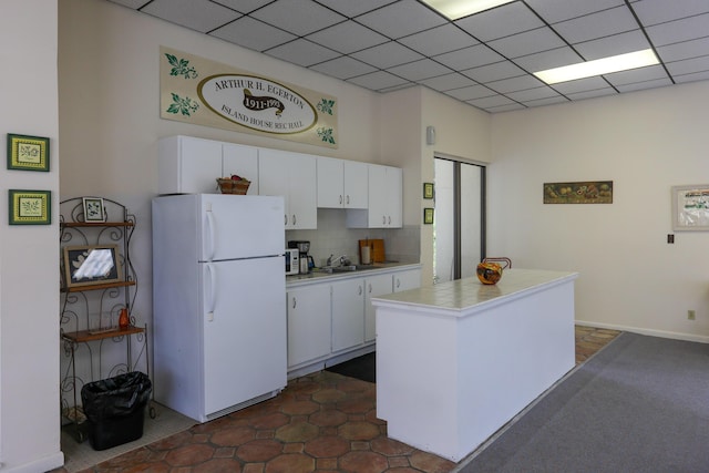 kitchen with a drop ceiling, decorative backsplash, freestanding refrigerator, a sink, and white cabinetry