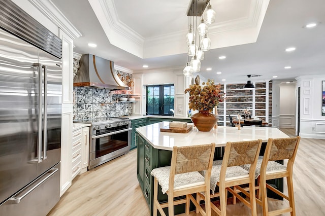 kitchen featuring wall chimney exhaust hood, a raised ceiling, high end appliances, green cabinetry, and a center island