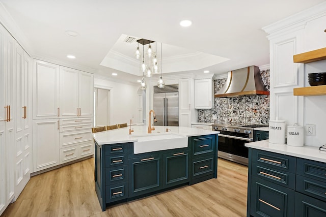 kitchen featuring high end appliances, white cabinetry, wall chimney exhaust hood, a tray ceiling, and ornamental molding