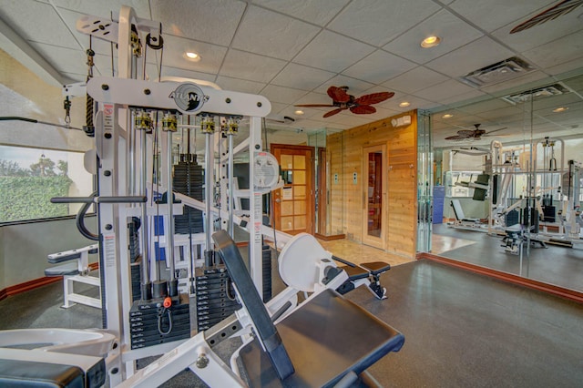 gym with a paneled ceiling, wooden walls, and ceiling fan