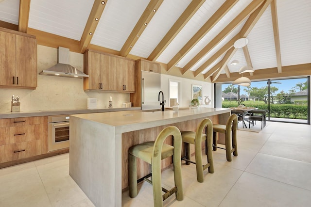 kitchen with appliances with stainless steel finishes, a kitchen bar, beam ceiling, a kitchen island with sink, and wall chimney range hood