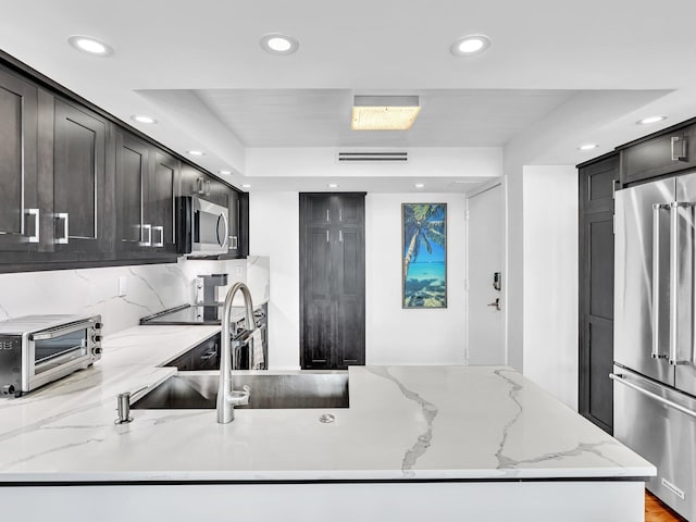 kitchen with a raised ceiling, decorative backsplash, light stone counters, kitchen peninsula, and stainless steel appliances