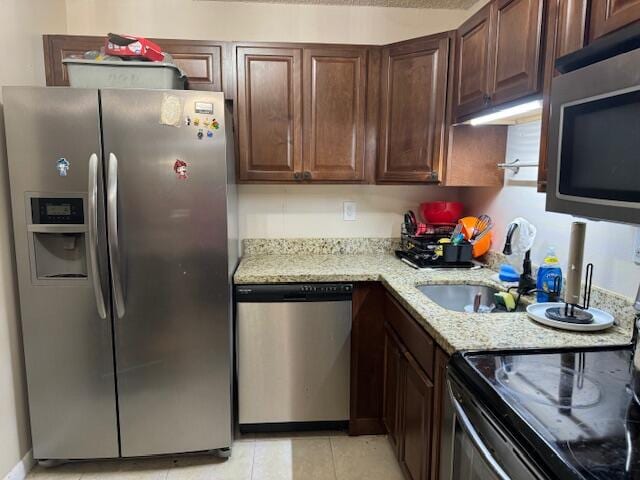 kitchen with dark brown cabinetry, light tile patterned floors, sink, stainless steel appliances, and light stone countertops