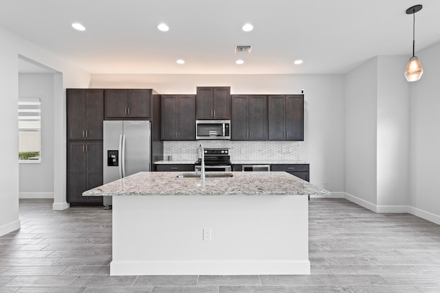 kitchen featuring appliances with stainless steel finishes, light stone counters, an island with sink, decorative light fixtures, and sink