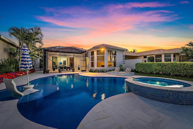 pool at dusk featuring glass enclosure, an in ground hot tub, and a patio