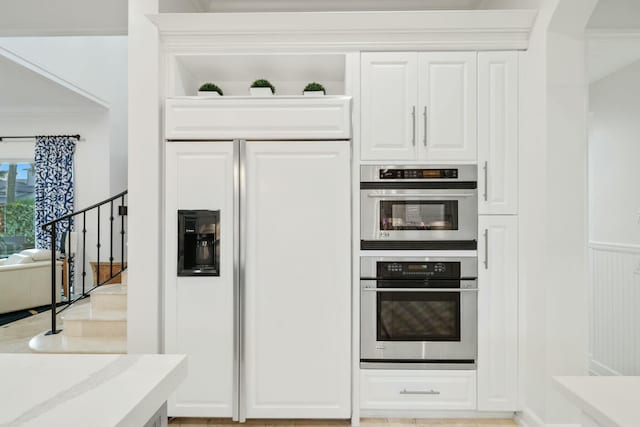 kitchen with light stone countertops, ornamental molding, stainless steel double oven, paneled built in refrigerator, and white cabinetry