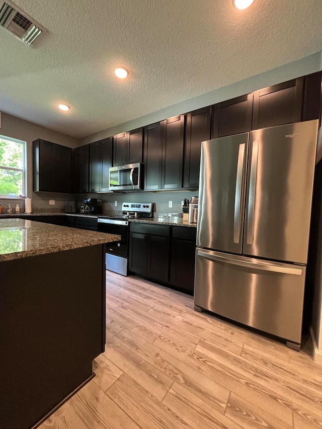 kitchen featuring a textured ceiling, stainless steel appliances, light hardwood / wood-style floors, and dark stone countertops