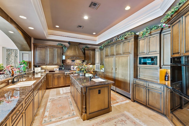 kitchen featuring dark stone counters, custom exhaust hood, a raised ceiling, sink, and a kitchen island