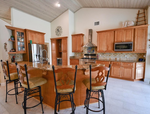 kitchen featuring a kitchen island with sink, stainless steel appliances, a breakfast bar, wooden ceiling, and wall chimney range hood