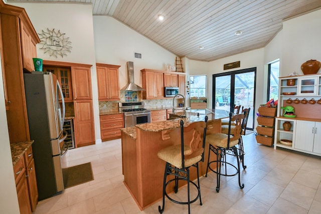 kitchen with light stone countertops, wall chimney exhaust hood, wooden ceiling, appliances with stainless steel finishes, and a center island with sink