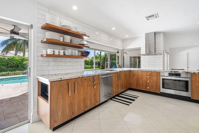 kitchen featuring appliances with stainless steel finishes, light stone counters, wall chimney exhaust hood, sink, and light tile patterned flooring