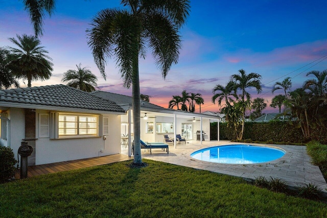 pool at dusk with a patio, ceiling fan, and a lawn