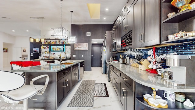 kitchen with light stone counters, dark brown cabinets, decorative light fixtures, a center island with sink, and appliances with stainless steel finishes