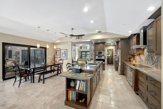 kitchen featuring pendant lighting, wall chimney range hood, a center island with sink, appliances with stainless steel finishes, and light stone countertops