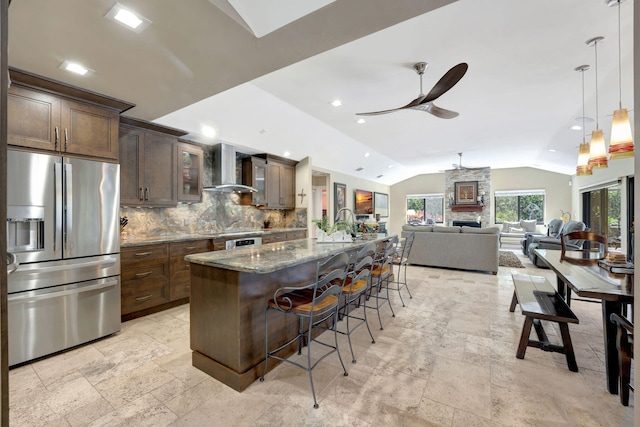 kitchen with an island with sink, wall chimney exhaust hood, stainless steel refrigerator with ice dispenser, a kitchen breakfast bar, and vaulted ceiling