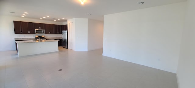 kitchen with light tile patterned flooring, stainless steel appliances, dark brown cabinets, and an island with sink