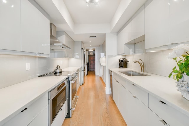 kitchen with white cabinetry, stainless steel appliances, wall chimney exhaust hood, sink, and light hardwood / wood-style flooring