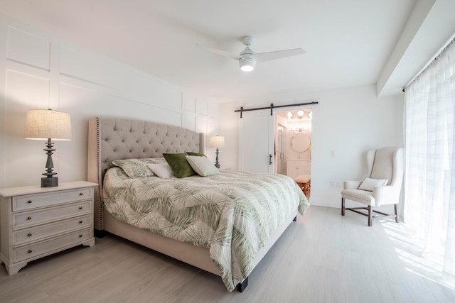 bedroom with a barn door, connected bathroom, light wood-type flooring, and ceiling fan