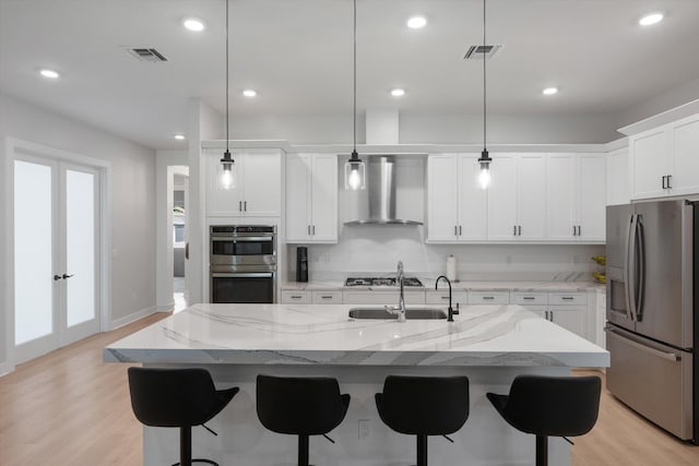 kitchen with light hardwood / wood-style floors, wall chimney range hood, a center island with sink, decorative light fixtures, and stainless steel appliances