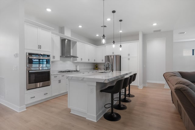 kitchen with light hardwood / wood-style flooring, a kitchen island with sink, wall chimney exhaust hood, white cabinetry, and appliances with stainless steel finishes