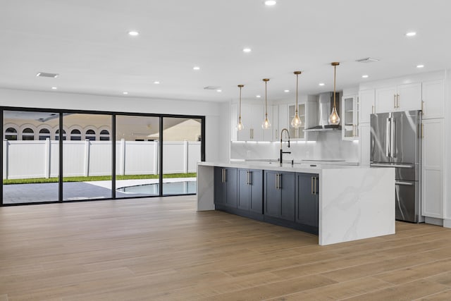 kitchen featuring an island with sink, freestanding refrigerator, wall chimney exhaust hood, white cabinetry, and recessed lighting