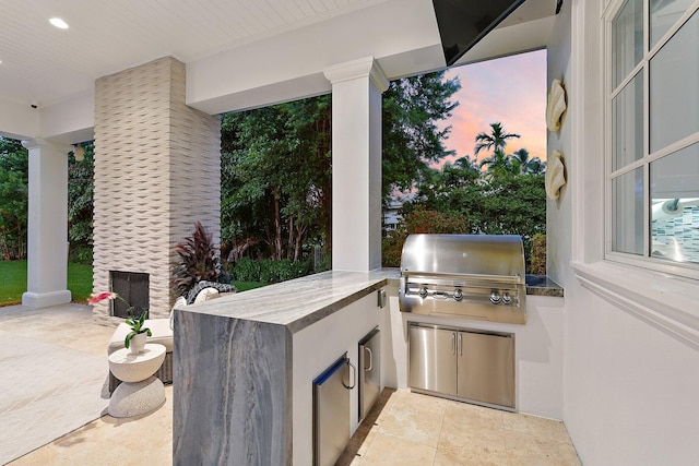 patio terrace at dusk featuring exterior kitchen, grilling area, and an outdoor stone fireplace