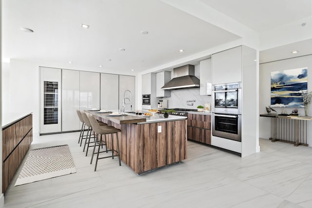 kitchen with a center island with sink, white cabinets, tasteful backsplash, wall chimney range hood, and appliances with stainless steel finishes