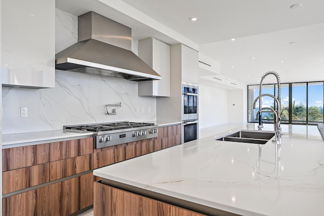 kitchen featuring stainless steel appliances, sink, light stone counters, wall chimney range hood, and decorative backsplash