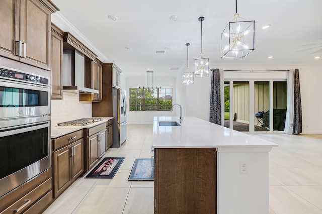 kitchen with appliances with stainless steel finishes, decorative light fixtures, a kitchen island with sink, sink, and wall chimney range hood