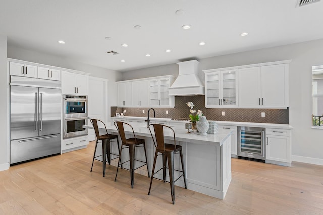 kitchen featuring a center island with sink, beverage cooler, appliances with stainless steel finishes, premium range hood, and light hardwood / wood-style floors