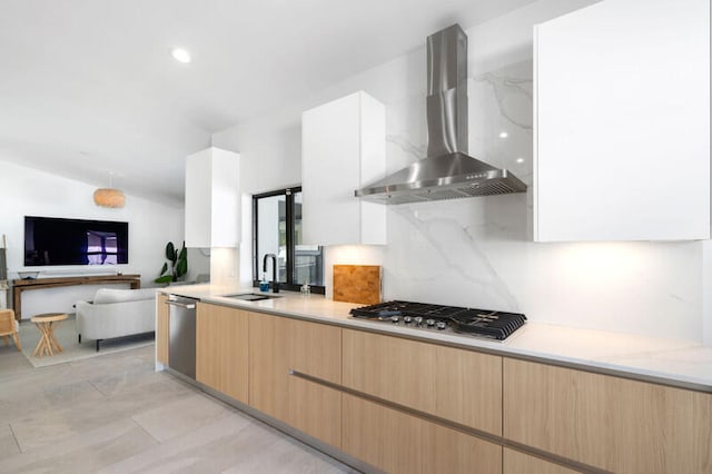 kitchen featuring wall chimney range hood, white cabinets, appliances with stainless steel finishes, light stone countertops, and sink