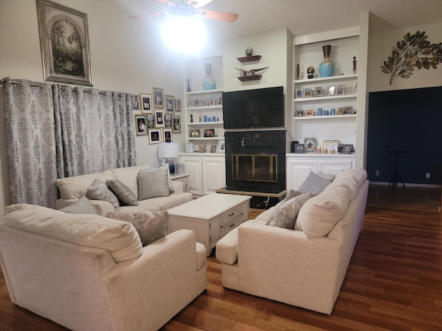 living room with ceiling fan, a premium fireplace, built in shelves, and dark wood-type flooring