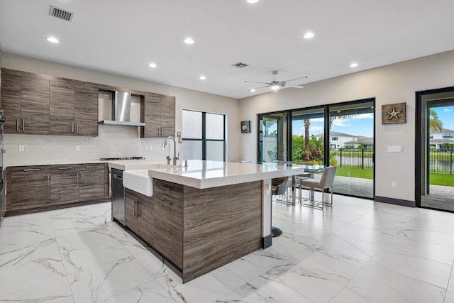 kitchen with wall chimney exhaust hood, an island with sink, backsplash, sink, and ceiling fan