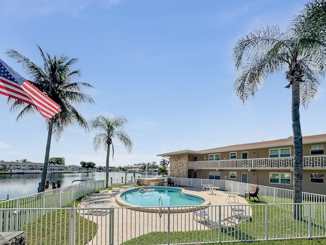 community pool with a patio, a yard, a water view, and fence