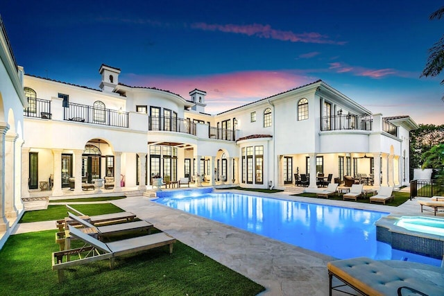 back house at dusk featuring a swimming pool with hot tub, a patio, and a balcony