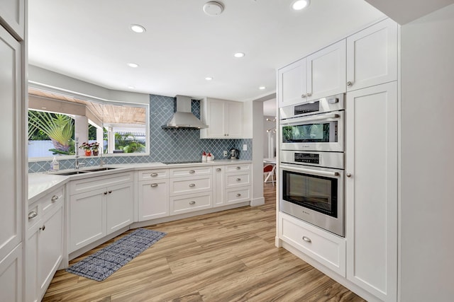 kitchen with wall chimney range hood, white cabinetry, double oven, light hardwood / wood-style floors, and decorative backsplash