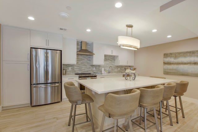 kitchen featuring wall chimney range hood, hanging light fixtures, white cabinetry, appliances with stainless steel finishes, and light hardwood / wood-style floors