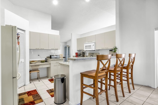 kitchen with light tile patterned flooring, kitchen peninsula, white appliances, backsplash, and a kitchen breakfast bar