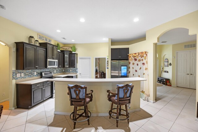 kitchen featuring tasteful backsplash, dark brown cabinets, stainless steel appliances, light tile patterned floors, and a center island with sink