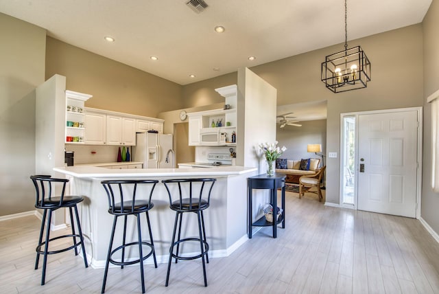 kitchen with white appliances, ceiling fan with notable chandelier, kitchen peninsula, light wood-type flooring, and white cabinetry