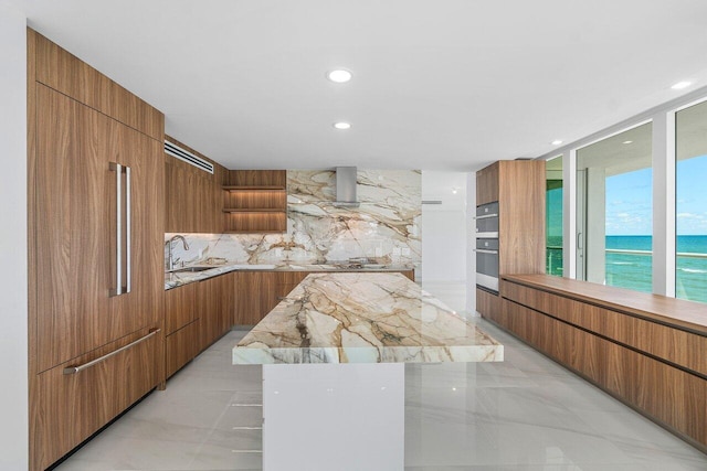 kitchen with a spacious island, wall chimney exhaust hood, sink, a water view, and backsplash