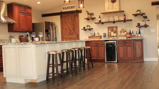 kitchen featuring light stone counters, wall chimney exhaust hood, vaulted ceiling, a center island with sink, and a breakfast bar area