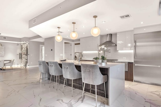 kitchen featuring pendant lighting, a kitchen island with sink, wall chimney range hood, white cabinetry, and stainless steel built in refrigerator