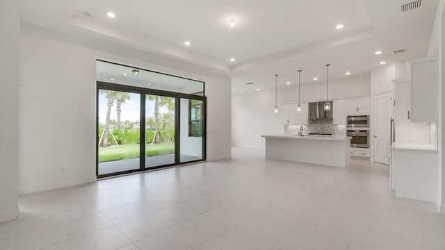 interior space with wall chimney range hood, pendant lighting, a raised ceiling, an island with sink, and white cabinetry