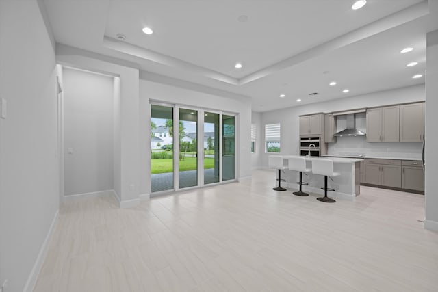 kitchen with an island with sink, gray cabinetry, wall chimney range hood, a tray ceiling, and a kitchen breakfast bar